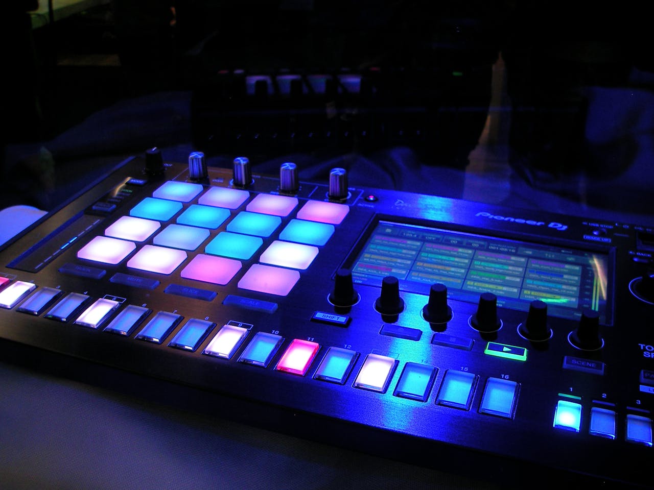 our-services-2 Close-up of an illuminated DJ mixer with vibrant LED pads in a dark studio environment.